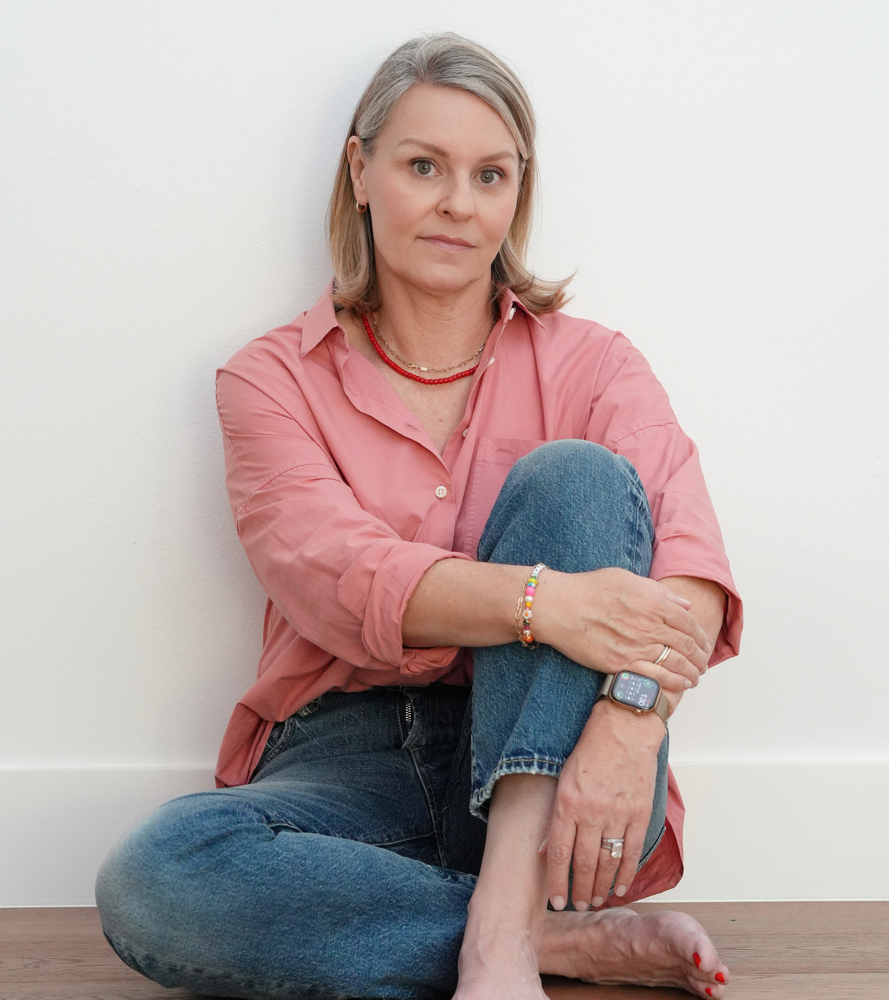 Patrina Odette sitting on a wood floor against a white wall in a coral shirt and jeans